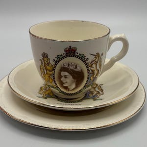 May include: A cream-coloured teacup, saucer, and plate set. The teacup and saucer feature a portrait of a woman wearing a crown, surrounded by ornate designs and the word "CORONATION". The set has gold trim.