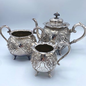 May include: A silver tea set with a teapot, creamer, and sugar bowl. Each piece is ornately decorated with floral designs.
