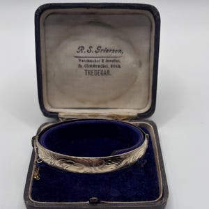 Könnte beinhalten: Ein silberfarbener Metallarmreif mit eingraviertem Blumenmuster und einer kleinen Sicherheitskette. Das Armband befindet sich in einer mit dunkelblauem Samt ausgekleideten Schmuckschatulle, auf deren Innendeckel der Text R.S. Grierson, Watchmaker & Jeweller, 23, Commercial Road, Tredegar aufgedruckt ist.