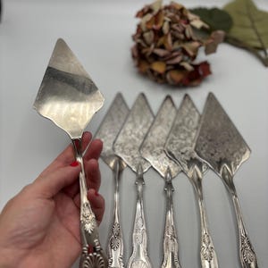 Könnte beinhalten: Eine Sammlung von silbernen Vintage-Kuchenservierern mit verzierten Griffen und dreieckigen Klingen. Die Servierer sind auf einer weißen Oberfläche angeordnet, mit einem dekorativen Blumenarrangement im Hintergrund. Die Kuchenservierer sind wahrscheinlich aus versilbertem Metall.