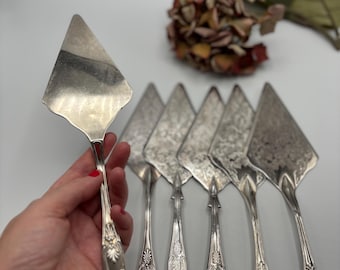 Könnte beinhalten: Eine Sammlung von silbernen Vintage-Kuchenservierern mit verzierten Griffen und dreieckigen Klingen. Die Servierer sind auf einer weißen Oberfläche angeordnet, mit einem dekorativen Blumenarrangement im Hintergrund. Die Kuchenservierer sind wahrscheinlich aus versilbertem Metall.