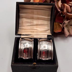 Könnte beinhalten: Zwei silberne Serviettenringe mit ovalem Design in einer schwarzen Box mit Samtinnenfutter. Die Ringe haben ein strukturiertes Muster. Getrocknete Blumen bilden den Hintergrund.