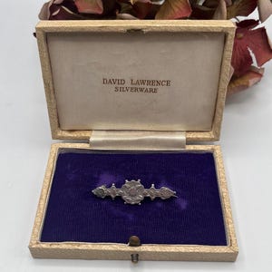 May include: A silver brooch with a floral design in a beige box with the text "DAVID LAWRENCE SILVERWARE" on the lid.