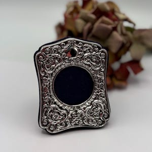 May include: A small, ornate silver picture frame with a dark blue interior. The frame has a rectangular shape with rounded corners and is decorated with intricate floral and scrollwork patterns. The background is blurred, with dried flowers.