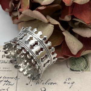 Puede incluir: Anillo de servilleta de plata con un diseño decorativo de calado. El anillo está hecho de dos bandas con un patrón repetido de diamantes y cuadrados.