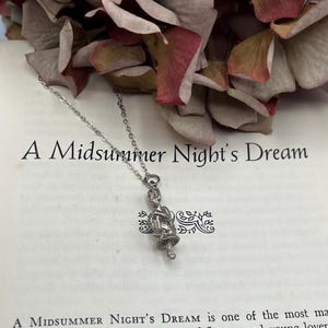 May include: A silver necklace with a charm of a woman holding a flower. The charm is detailed and intricate. The necklace is on a book with the text "A Midsummer Night's Dream".