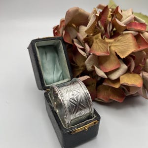 Puede incluir: Un servilletero plateado con un patrón geométrico grabado, exhibido en una caja negra vintage con interior azul claro. El anillo se encuentra sobre un fondo de flores secas multicolores.