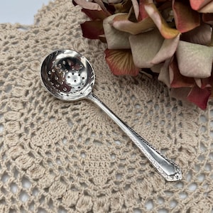 Peut inclure: Une cuillère à thé en argent avec un bol perforé et un manche orné. La cuillère repose sur un napperon en crochet.