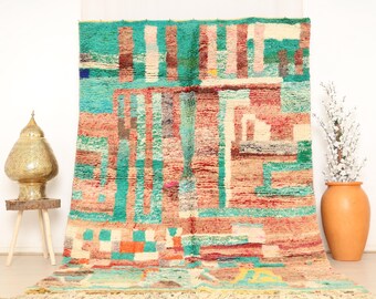 Puede incluir: Una colorida alfombra rectangular con un diseño geométrico. La alfombra presenta una mezcla de colores turquesa, rosa, naranja y crema. Tiene flecos en el borde inferior y se muestra sobre un fondo blanco.