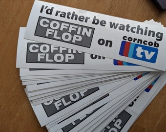Puede incluir: Una pila de pegatinas blancas con el texto "I'd rather be watching COFFIN FLOP on corncob tv" impreso en negro. Las pegatinas están impresas con un logotipo en blanco y negro de "COFFIN FLOP" y un logotipo azul, rojo y blanco de "corncob tv".