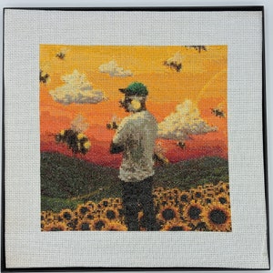 May include: A framed cross-stitch artwork depicting a person standing in a field of sunflowers with a sunset in the background. The person is wearing a green baseball cap and a white shirt. There are bees flying around the person and the sunflowers.