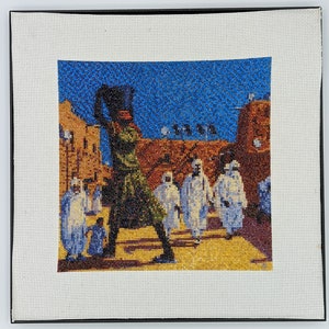 May include: Needlepoint artwork depicting a figure in green and black attire, holding an object aloft, set against a backdrop of a town with figures in white robes. The sky is blue, and the artwork is framed in black.