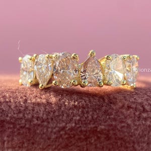 May include: A gold ring with multiple clear, pear-shaped gemstones. The ring is set on a pink velvet surface, with a pink background. The ring is from the brand "Lionza Diamond".