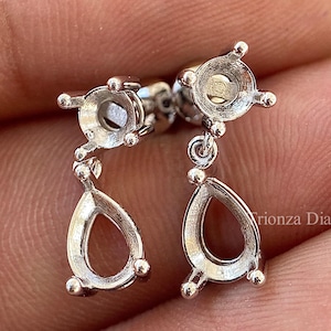 May include: A pair of silver-toned drop earrings. Each earring features a round top with a setting for a gemstone and a teardrop-shaped dangle. The earrings are displayed against a neutral background.