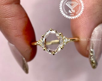 May include: A gold ring with a large, clear, oval-cut gemstone. The ring has smaller accent stones on either side of the main stone. The text "Trionza Diamond" is visible on the ring's band. The ring is held up by a person's fingers.