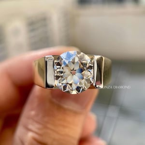 May include: A gold ring with a large, round, clear diamond. The diamond is faceted, reflecting light. The ring band is thick and rectangular. The text "ONZA DIAMOND" is visible on the ring.