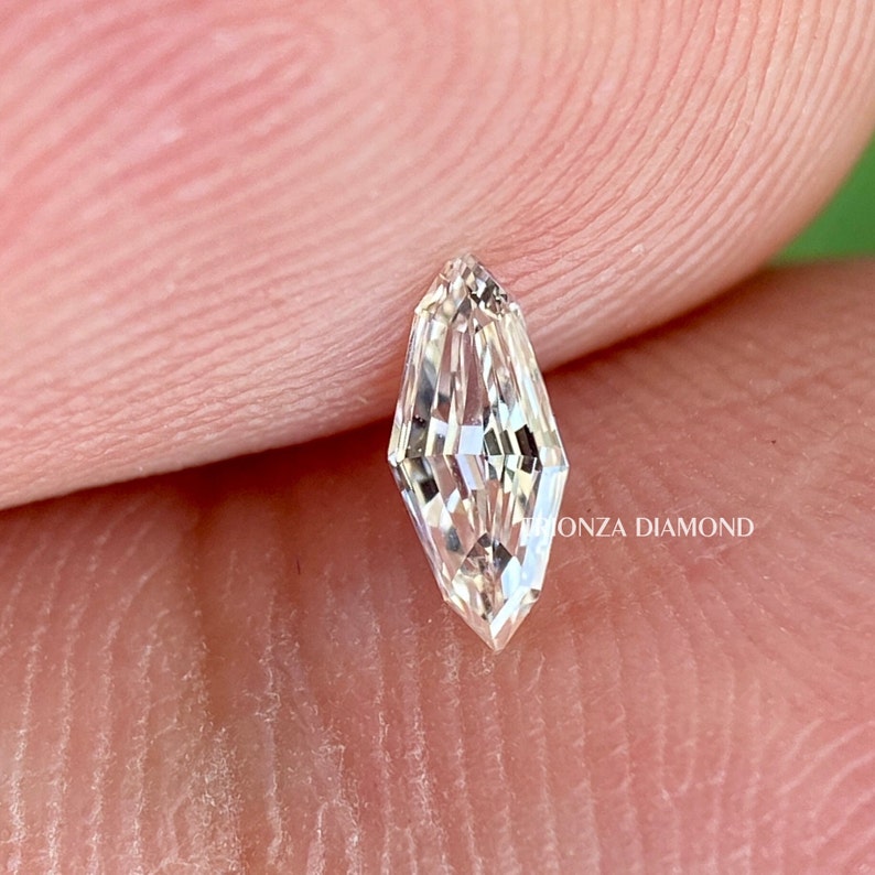 May include: A clear, faceted, elongated diamond-shaped gemstone with a pointed tip. The gemstone is set against a light pink background.