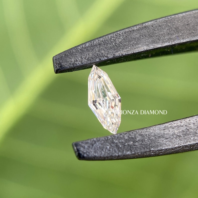 May include: A clear, faceted, elongated diamond held by tweezers. The diamond has a rectangular shape with a pointed top and bottom. The diamond is held against a green background.