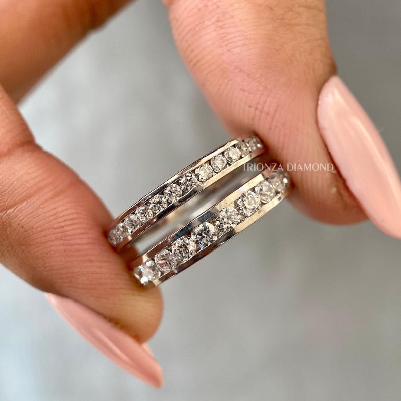 May include: A pair of silver eternity bands with a row of small diamonds set in each band. The bands are stacked on top of each other. The text "TRIONZA DIAMOND" is visible on the band.