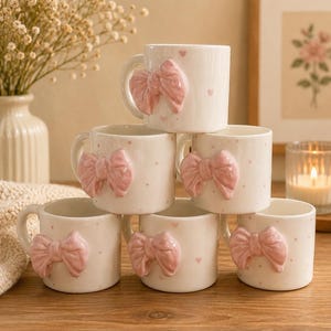 Puede incluir: Una pila de seis tazas de cerámica blanca, cada una con un lazo rosa tridimensional en relieve en la parte frontal. Las tazas están decoradas con un patrón de pequeños puntos de color rosa claro y diminutos corazones sobre la superficie.