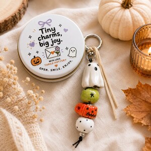May include: A round white metal tin with the text Tiny charms, big joy. WITH LOVE. TANCO POTTERY. OPEN. SMILE. ENJOY. and illustrations of a ghost, pumpkin, and envelope. A keychain features a white ghost, green star bead, orange pumpkin, and white polka dot bead.
