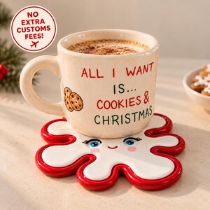Könnte beinhalten: Eine cremefarbene Keramiktasse mit dem Text ALL I WANT IS... COOKIES & CHRISTMAS und einer Keks-Illustration, die auf einem weiß-roten, wie Zuckerguss geformten Untersetzer mit lächelndem Gesicht, blauen Augen und rosa Wangen steht.