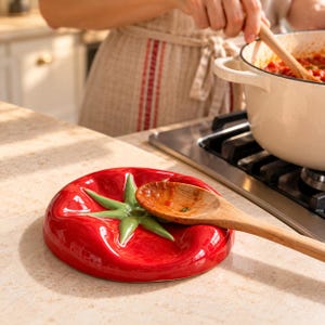 Puede incluir: Un reposacucharas de cerámica roja brillante con forma de tomate y un tallo verde en el centro. El plato redondo y cóncavo presenta una superficie lisa y reflectante, diseñada para sostener una cuchara de madera con salsa de tomate.