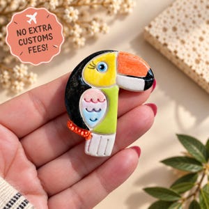 Peut inclure: Une broche en forme de toucan avec une tête et un dos noirs, un visage jaune vif avec un œil bleu, un grand bec orange et blanc, une poitrine vert clair, une aile à motifs roses et bleus et une queue blanche avec des accents orange.