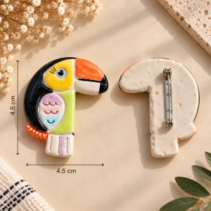 Puede incluir: Un broche de cerámica con forma de tucán que presenta un cuerpo negro, amarillo, naranja y verde claro con un detalle de ala rosa y azul. El broche mide 4,5 cm por 4,5 cm e incluye un alfiler de metal en la parte posterior.