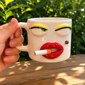 May include: A white ceramic mug featuring a three-dimensional face with raised yellow eyelids, green eyes, and glossy red lips. A small brown mole is visible on the cheek, and a cigarette with an orange filter is held between the lips.
