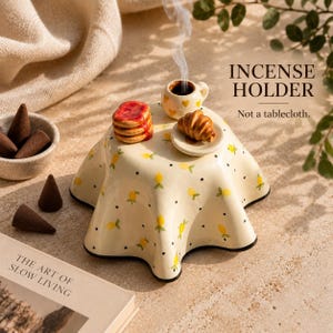 Può includere: Un porta incenso in ceramica a forma di tavolino rotondo con superficie color crema decorata da un motivo di limoni gialli e puntini neri. Include una tazzina e un piattino in miniatura con cibo. Testo: INCENSE HOLDER. Not a tablecloth.