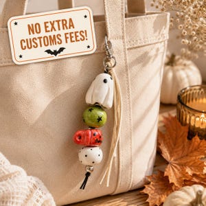 Op de afbeelding: Een beige canvas tas met een hangende bedel bestaande uit een witte spookkraal, een groene kraal met sterpatroon, een rode kraal met sterpatroon en een witte kraal met stippen. Een label aan de tas toont de tekst NO EXTRA CUSTOMS FEES! met een vleermuis.