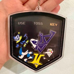 Könnte beinhalten: Ein schildförmiger Schlüsselanhänger aus klarem Acryl mit schwarzem Hintergrund und dem verpixelten Text USE, TOSS und KEY oben. Im Inneren befinden sich fünf bunte Charakter-Anhänger, darunter ein Narr, eine blau gekleidete Figur, ein Stern und eine weißgesichtige Figur.