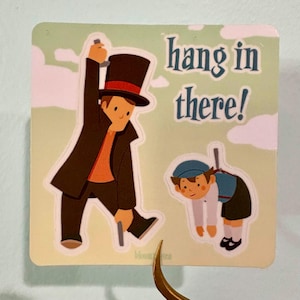 May include: A sticker with a cartoon illustration of two figures. One figure in a top hat and coat is holding a cane, while the other hangs from a pole. The text "hang in there!" is above the figures.