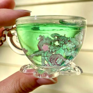 May include: A clear, miniature teacup keychain filled with green liquid and featuring anime-style characters. The teacup has a gold-colored chain and a decorative base. The characters are in shades of green, pink, and white.