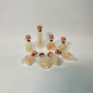 May include: A collection of small, clear glass bottles with cork stoppers. Each bottle contains a light yellow, fibrous material. The bottles vary in shape, including round, oval, and cylindrical designs. The arrangement is on a white surface.