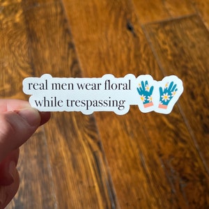 May include: White sticker with the text "real men wear floral while trespassing" and a pair of blue gardening gloves with white flowers printed on them.