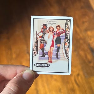 May include: A white sticker with a Clueless movie poster featuring three women in a staircase. The text on the poster reads "See, Cher, Popularity is a Hard Problem, Huh?" and "Clueless".