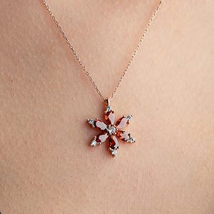 May include: A delicate necklace with a flower pendant. The pendant features a starburst design with red and white gemstones set in a gold-colored metal. The necklace is worn against a dark blue garment.