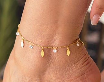 Puede incluir: Una tobillera dorada con un diseño de cadena delicada. La tobillera presenta pequeños dijes en forma de hoja y piedras preciosas claras y brillantes. La joya se lleva en un tobillo, mostrando los intrincados detalles y el estilo elegante.