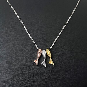May include: A silver necklace featuring three small, stylized fish pendants in rose gold, silver, and gold colors. The pendants are suspended from a delicate silver chain, displayed on a black mannequin.