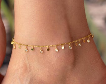 May include: A gold-colored anklet with dangling charms. The anklet features small, round, and teardrop-shaped charms, some with clear stones. The jewelry is worn on an ankle, with a hand and foot visible in the image.