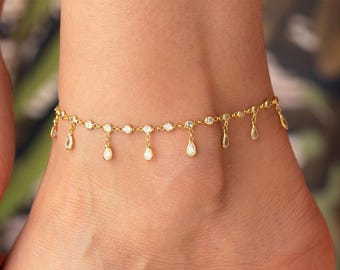 May include: A gold-colored anklet with a delicate design. The anklet features small, clear gemstones and teardrop-shaped charms that dangle from the chain. The jewelry is worn on a person's ankle.