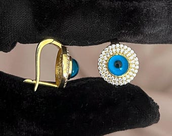 May include: A pair of gold-colored hoop earrings, each featuring a blue eye-shaped center stone surrounded by a halo of small, clear crystals. The earrings are displayed against a black background.