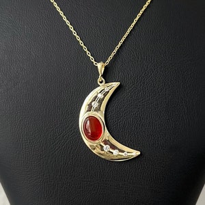 May include: A gold crescent moon pendant necklace with a red gemstone in the centre and small clear gemstones around the edge.