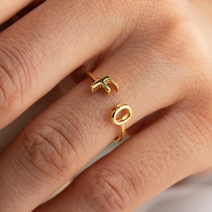 Puede incluir: Un anillo dorado con las letras "F" y "O" como diseño. El anillo es abierto, con las letras a ambos lados del dedo. El anillo está en el dedo de una persona.