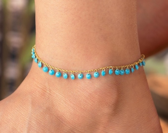 May include: A gold-tone anklet featuring small, turquoise beads suspended from a fine chain. The anklet is displayed on an ankle, highlighting the jewellery's design and colour contrast against the skin.