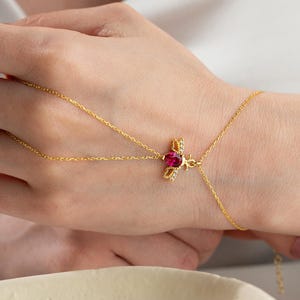 May include: A gold-colored hand chain with a central heart-shaped, ruby-red gemstone. The chain extends to the wrist and connects to a finger ring, creating a delicate jewelry piece. Small, clear gemstones surround the ruby.
