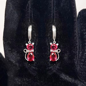 Puede incluir: Pendientes plateados con forma de gato y piedras preciosas rojas. Cada pendiente presenta un diseño de gato, con una cabeza de gema redonda y un cuerpo de gema ovalado. Los pendientes se muestran sobre un fondo de terciopelo negro.