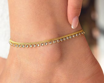 May include: A gold-colored anklet with small, clear, dangling charms. The delicate chain wraps around the ankle, showcasing the jewelry. The anklet is worn on a fair-skinned foot.